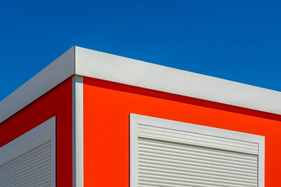 Minimalist mimari, parlak mavi gökyüzü arka planında kapanmış beyaz panjurlu bir kırmızı bina köşesini sergiliyor.